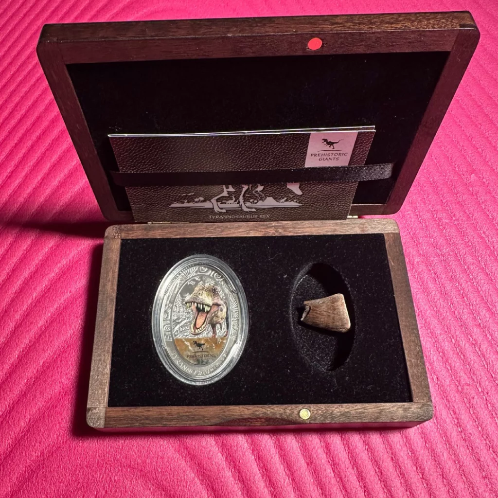 Wooden display box open to show a silver dinosaur coin and a small fossil tooth, labeled Prehistoric Giants.