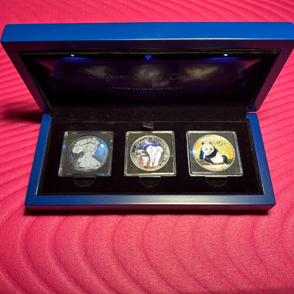 Blue presentation box opened to show three protective-cresentation coins, left coin with a detailed silhouetted figure, middle coin depicting wildlife elephants, right coin featuring a panda; all in clear plastic cases on a black insert against a red background.