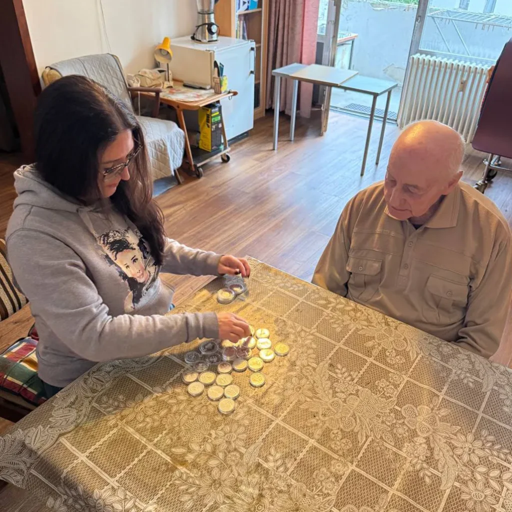 Woman and elderly man seated at a table, sorting or playing with a pile of coins on a patterned tablecloth in a bright room.