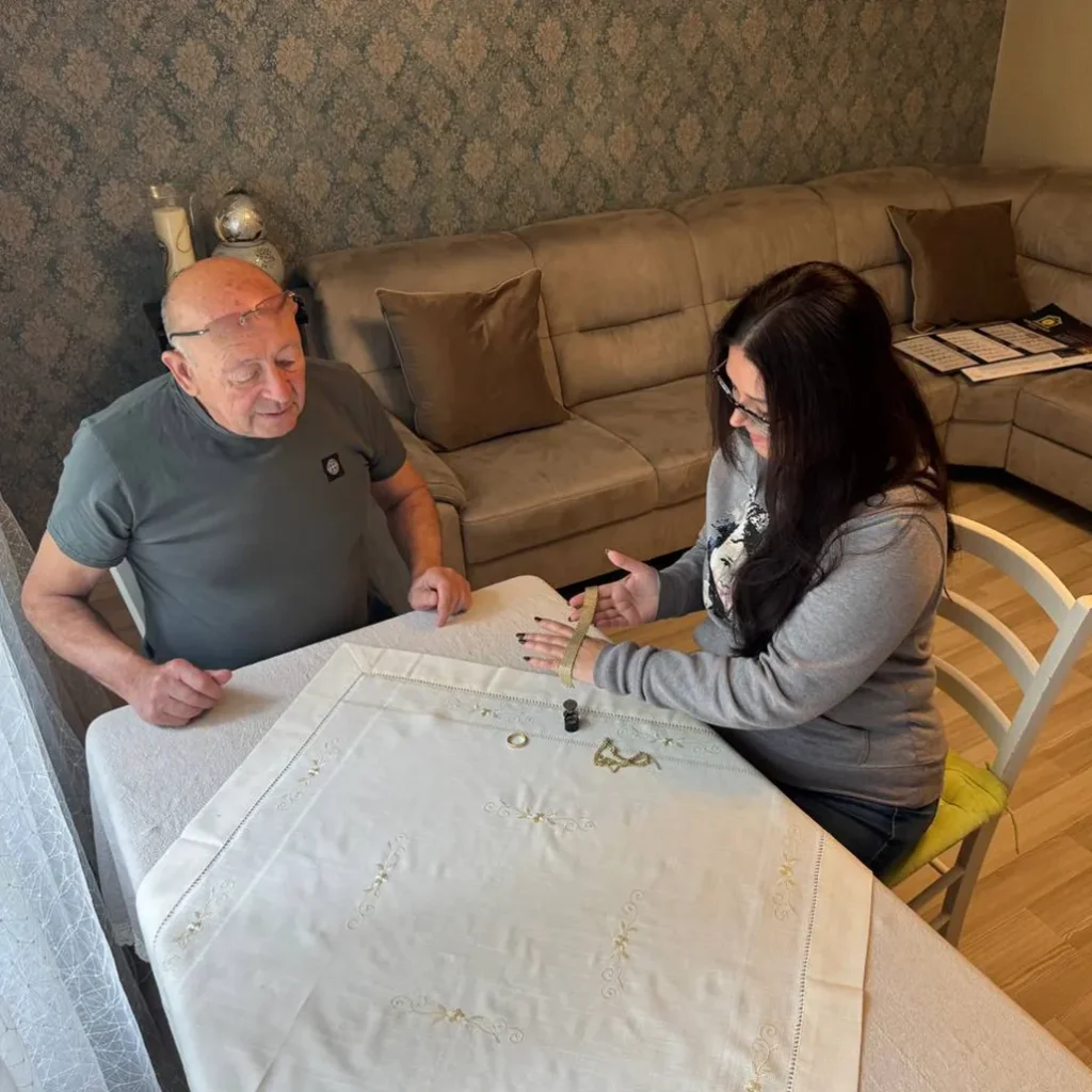 A man and a woman sit at a table measuring a light-colored embroidered tablecloth with a tape measure in a living room setting.