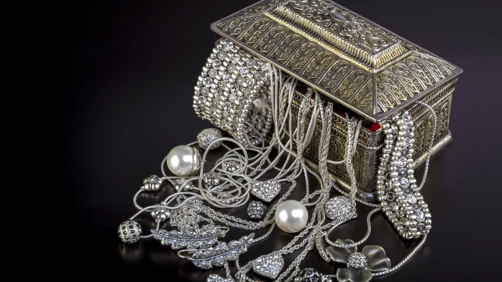 Open antique jewelry box with intricate metalwork spilling out silver necklaces, bracelets, and pearl strands onto a dark surface.