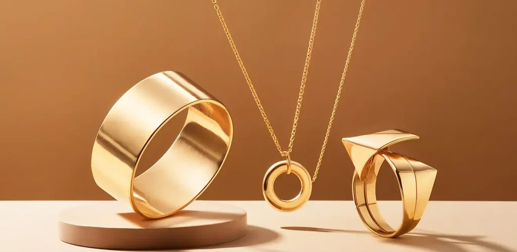 Gold jewelry set: a wide gold ring on a pedestal, a circular pendant necklace, and a modern geometric ring displayed on a warm brown backdrop.