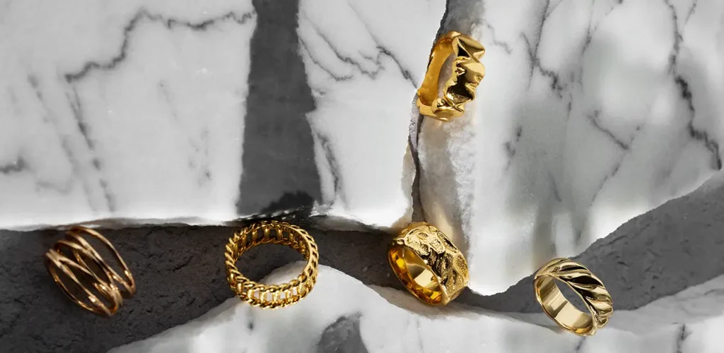 Gold rings arranged on a white marble surface with gray veining, featuring braided and twisted band designs fallowed by a broken wall shadowy edge.